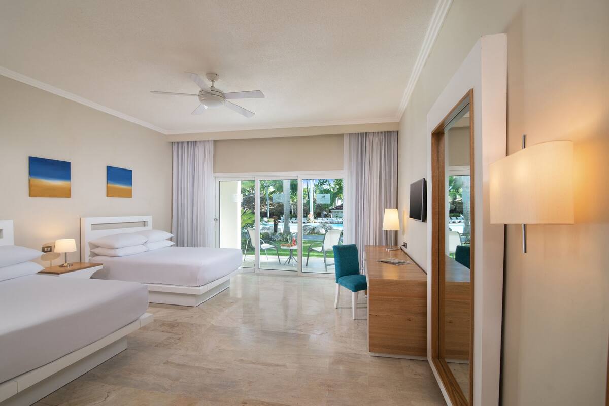 A hotel room with two beds, a desk, a chair, a TV, and a balcony with a view of the outdoors.
