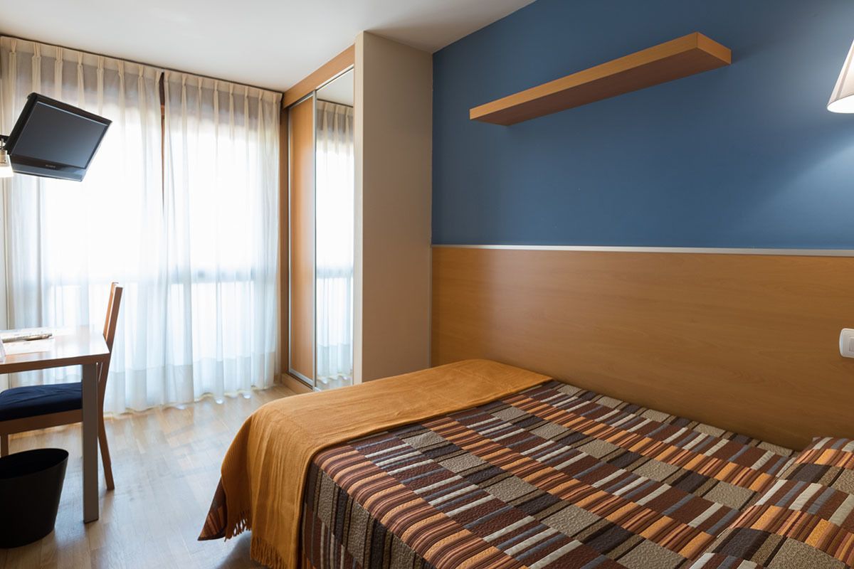 A hotel room with a bed, a desk with a chair, a TV mounted on the wall, and a window with curtains.