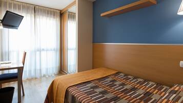A hotel room with a bed, a desk with a chair, a TV mounted on the wall, and a window with curtains.