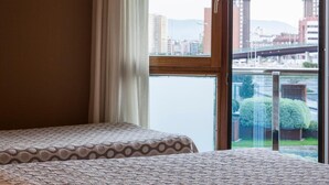 Habitación de hotel con dos camas, un ventanal con vistas a un paisaje urbano y un puente, y una lámpara a la izquierda.