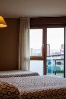 Habitación de hotel con dos camas, un ventanal con vistas a un paisaje urbano y un puente, y una lámpara a la izquierda.