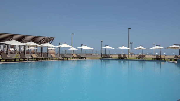 Una piscina con sillas de descanso y sombrillas, rodeada por una zona techada con vista a la playa.
