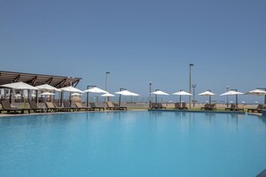 Una piscina con sillas de descanso y sombrillas, rodeada por una zona techada con vista a la playa.