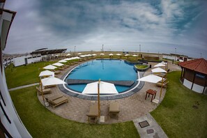 Una piscina circular con sillones de descanso y sombrillas, rodeada de césped y un edificio con techo rojo.