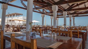 Un área de comedor al aire libre con mesas y sillas de madera, sombrillas blancas y vista al océano.