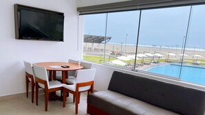 Una habitación con televisión, una mesa redonda con sillas, un sofá y un ventanal con vista a una piscina y una zona de playa.