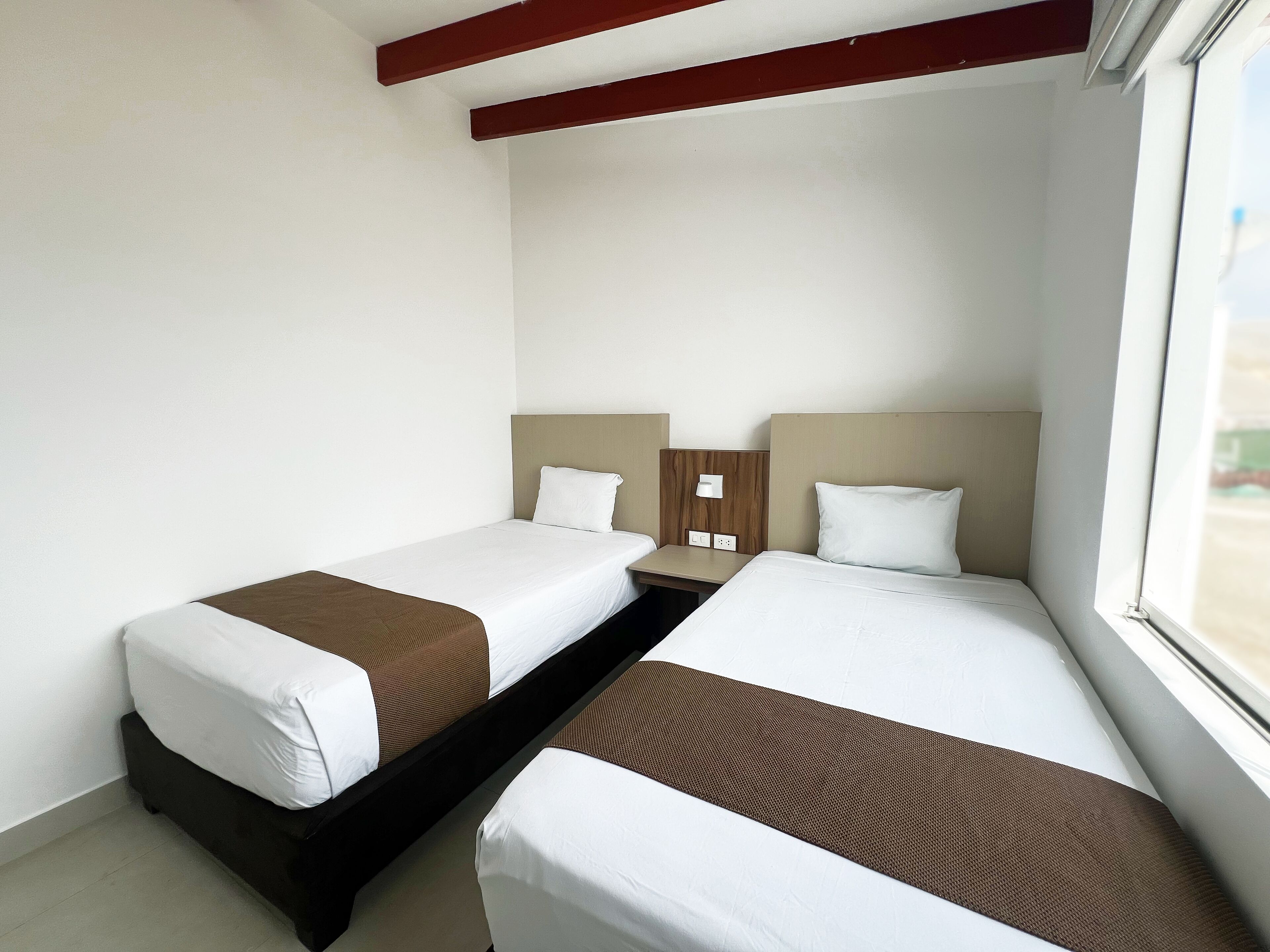Ein Hotelzimmer mit zwei Einzelbetten, einem Nachttisch, einem Fenster und Holzbalken an der Decke.