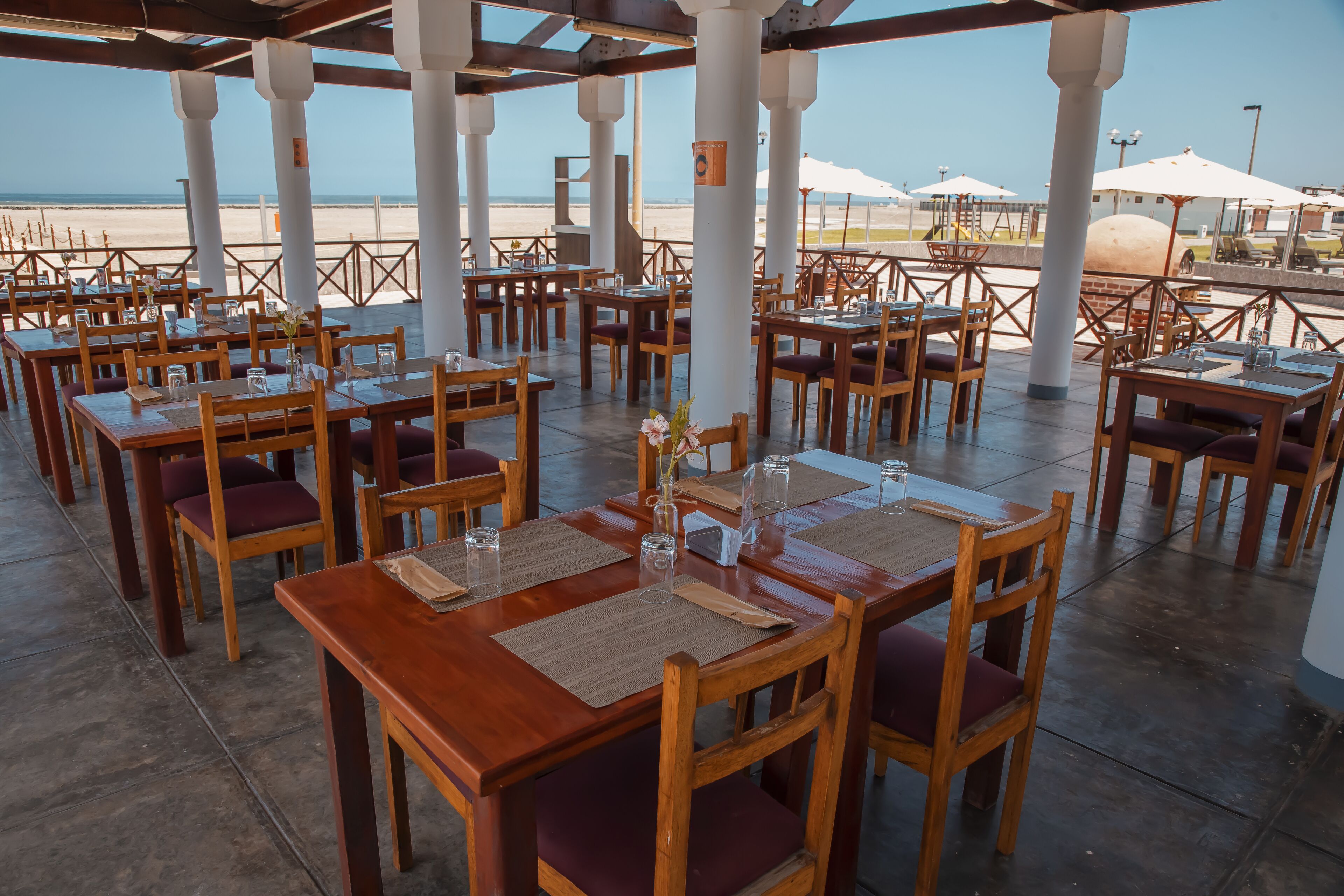 Restaurante ao ar livre com mesas e cadeiras de madeira, dispostas em fileiras, com vista para uma praia.