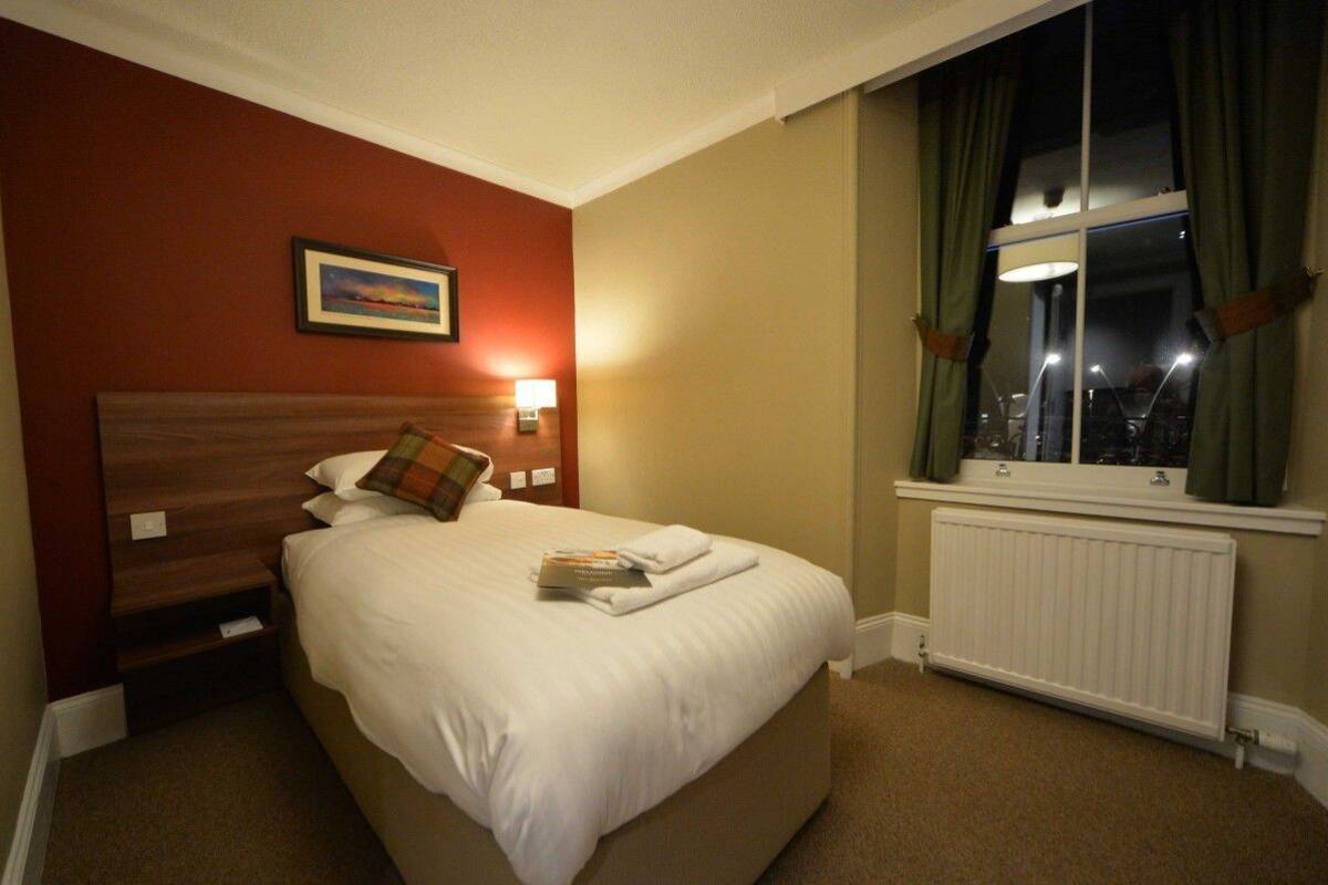 Ein Hotelzimmer mit einem großen Bett, einem Nachttisch mit Lampe, einem Fenster mit Vorhängen und einem gerahmten Bild an der Wand.