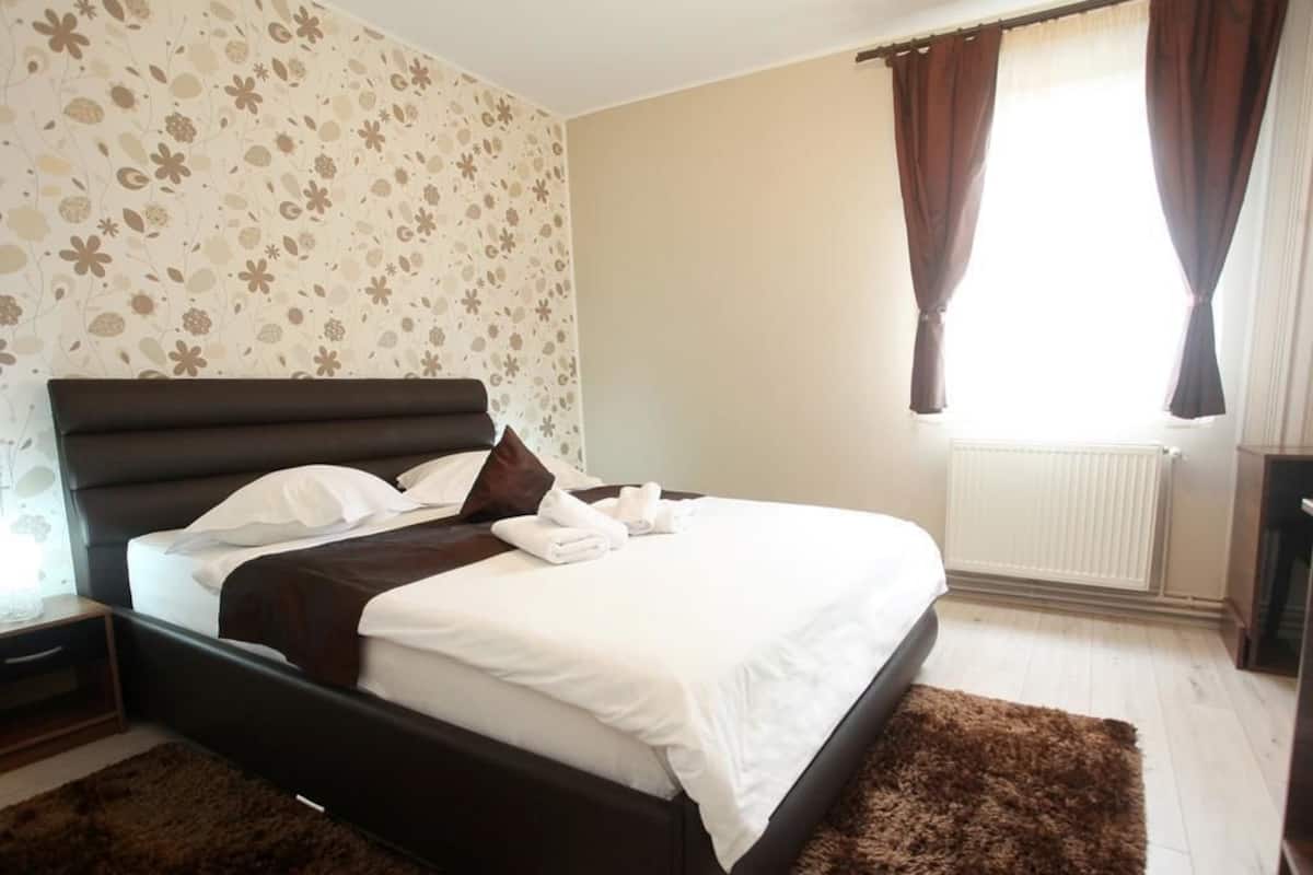 A neatly made bed with white linens and brown pillows, a dark headboard, and a brown rug on a light wooden floor.