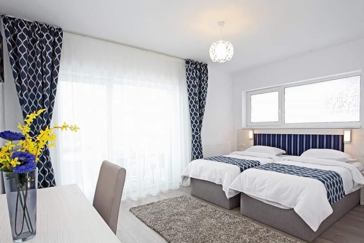 A modern hotel room with two beds, a dining area, and large windows.