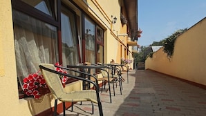 A corridor with wicker chairs and tables, a window with red flowers, and a wall with climbing plants. - Pension Mora Cluj Napoca (Cluj-Napoca)