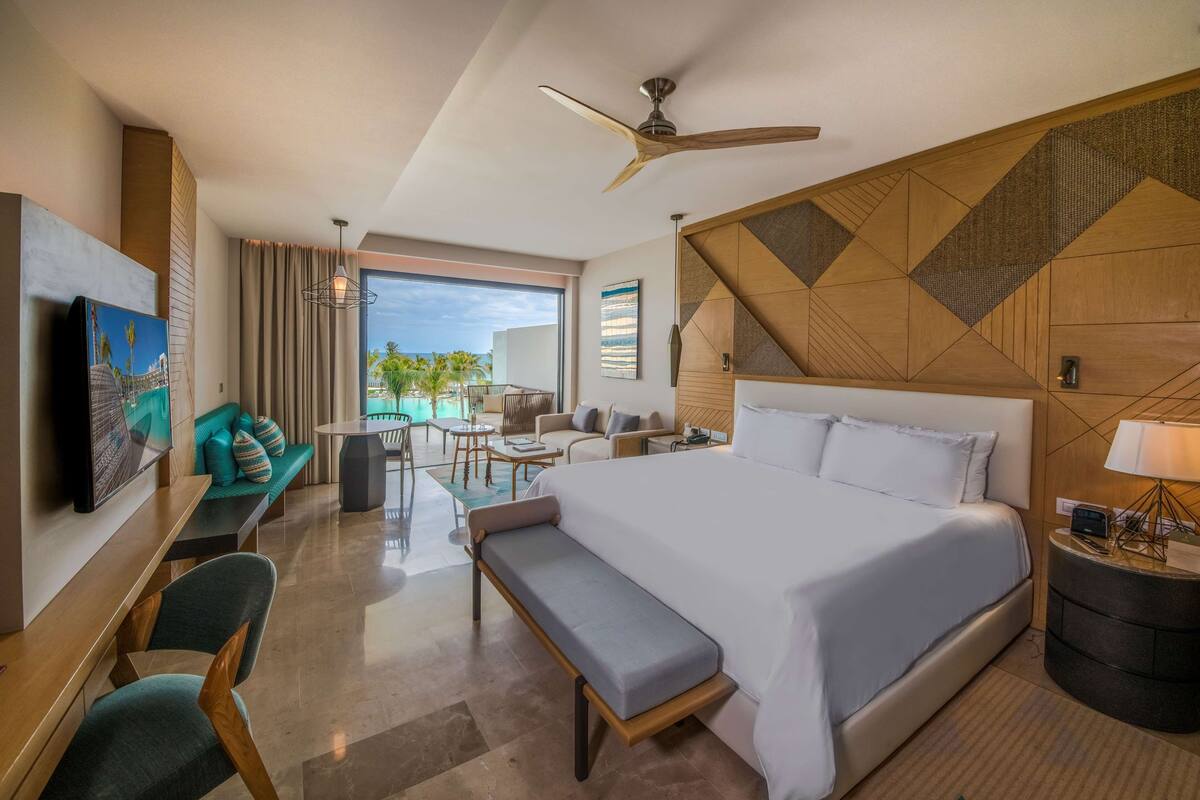 A modern hotel room with a large bed, a desk, a bench, a dining area, and a view of the outdoors.