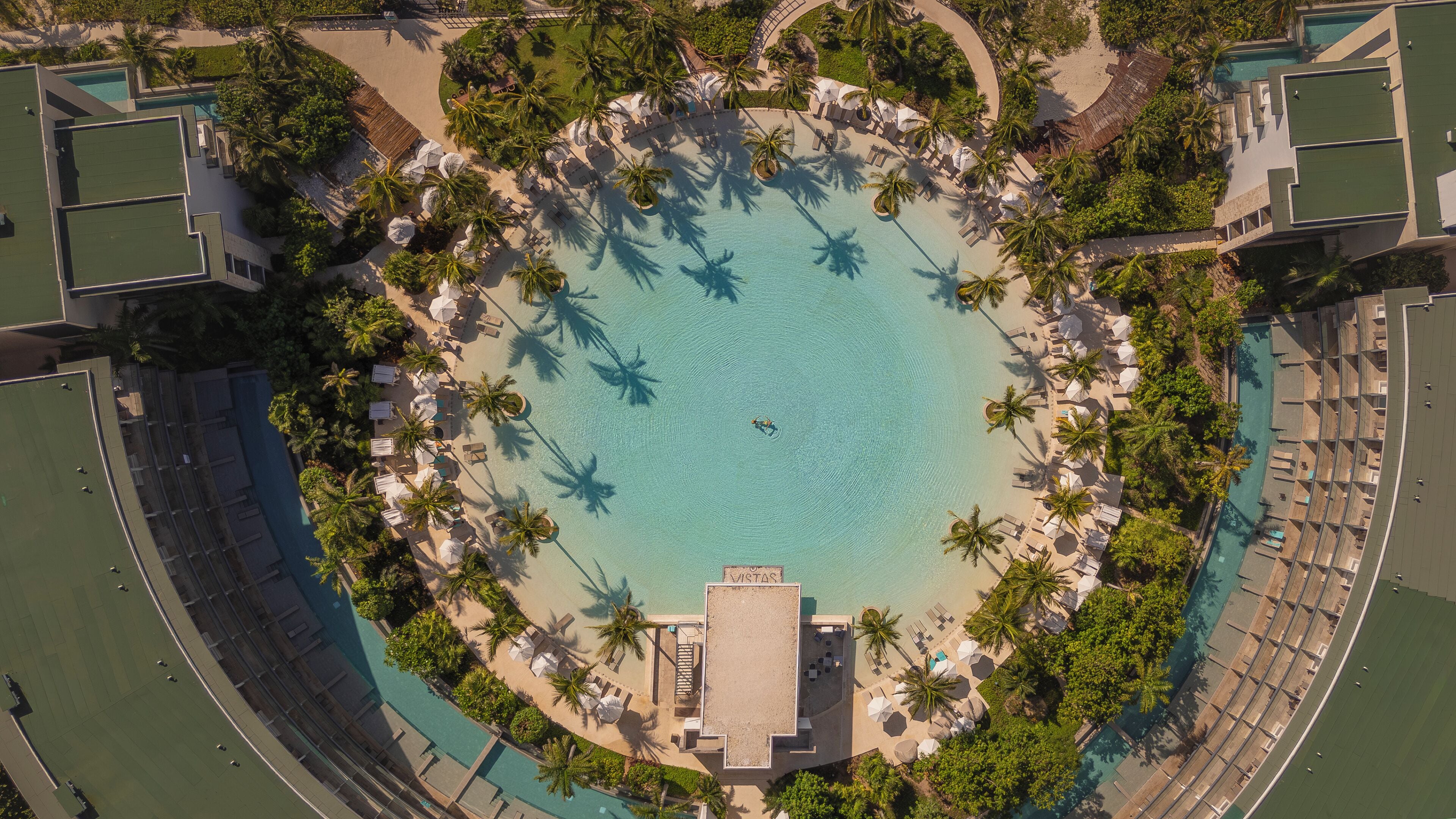 An aerial view of a resort with a central circular pool surrounded by palm trees and lounge areas.