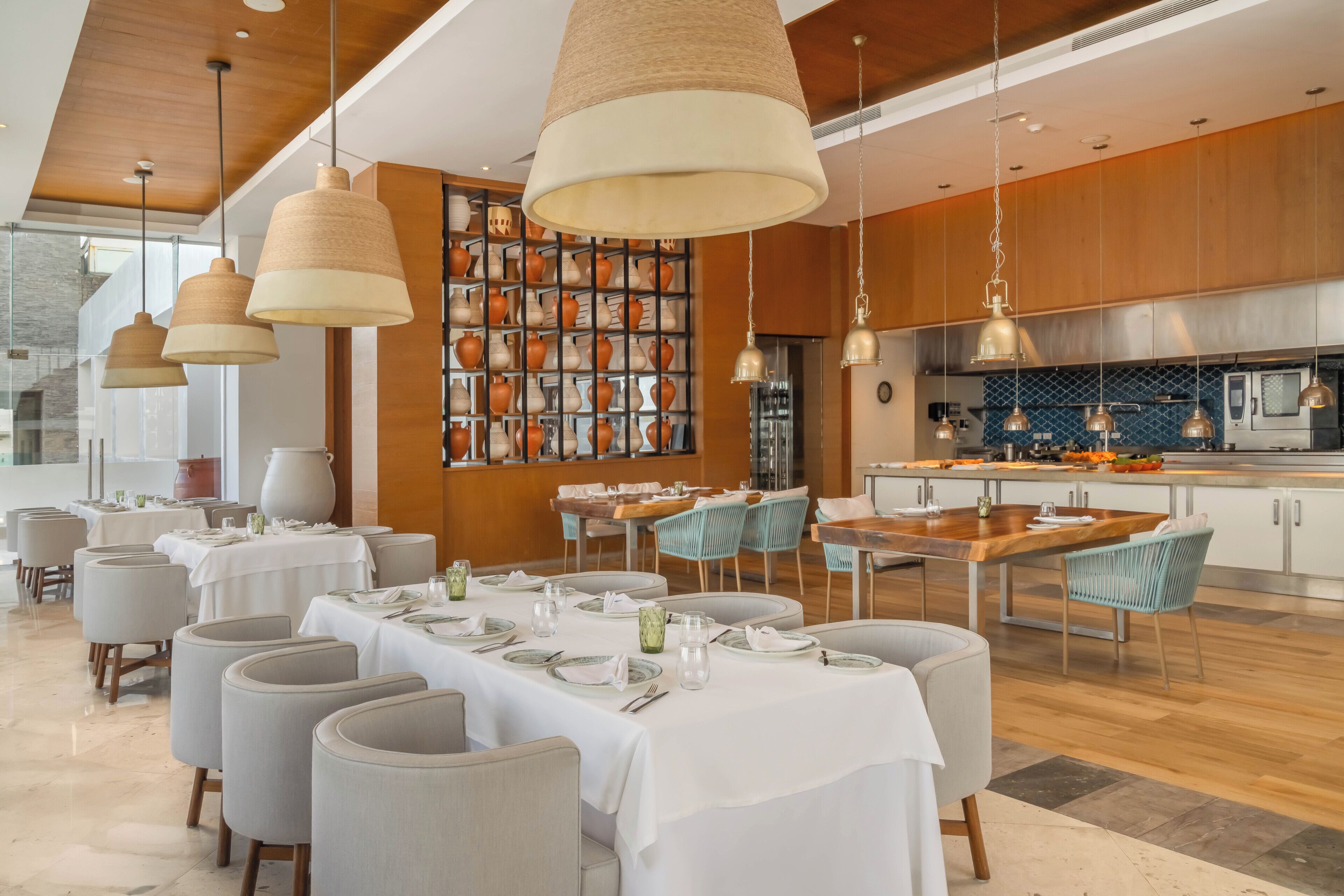 A modern dining area with multiple tables set for guests, featuring a mix of round and rectangular tables, light grey upholstered chairs, and hanging pendant lights.