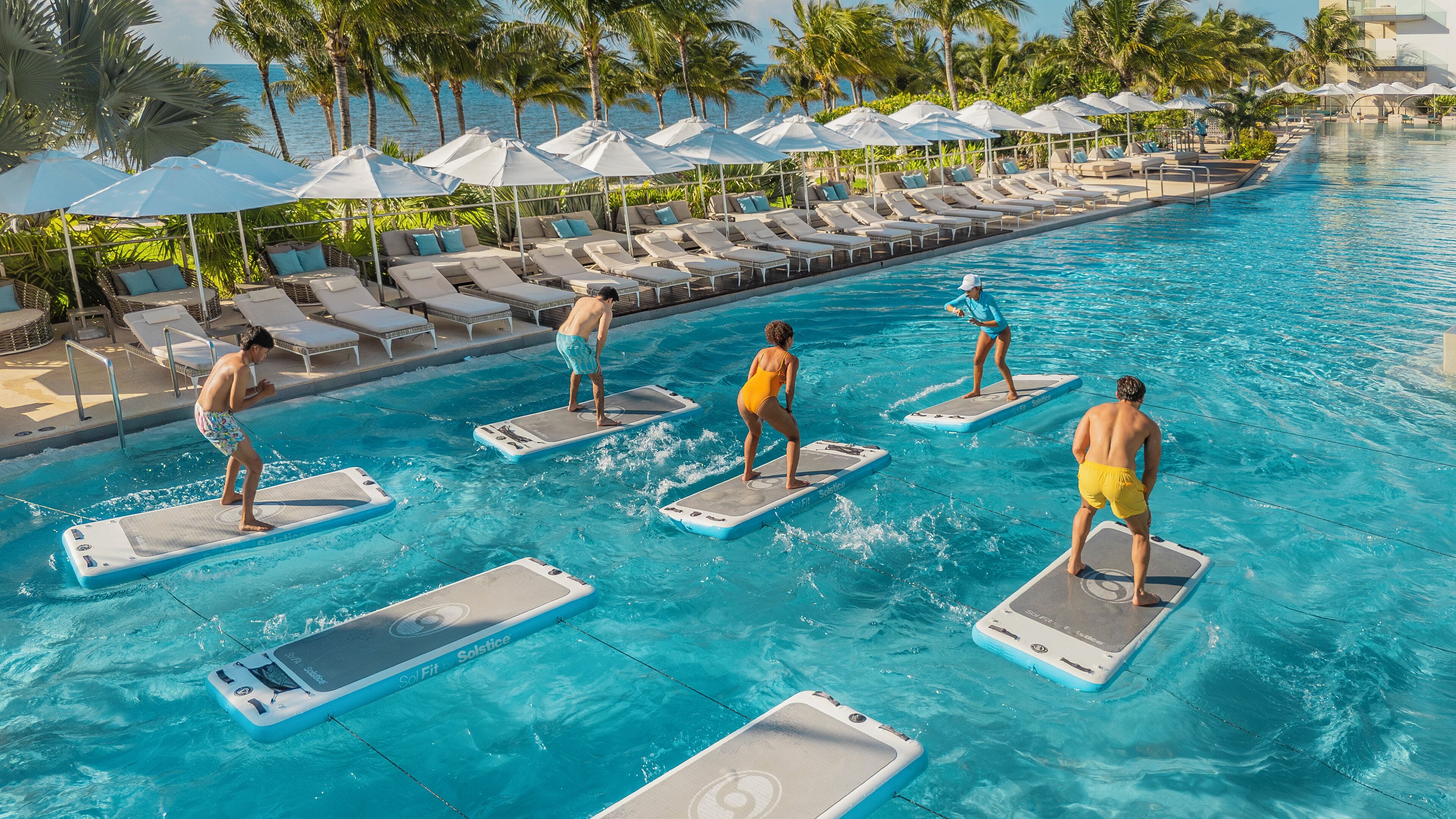 Une piscine avec des chaises longues et des parasols, et des personnes utilisant des bouées ou d’autres dispositifs de flottaison.