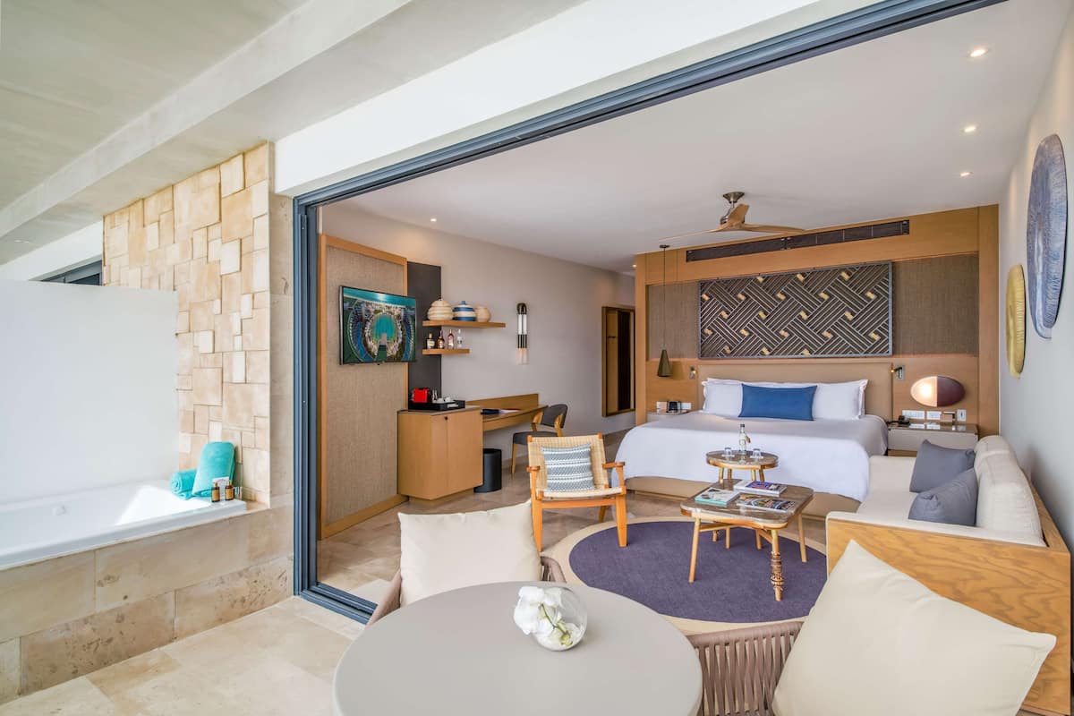 A modern hotel room with a large bed, a desk, a sofa, a round table, and a bathtub.