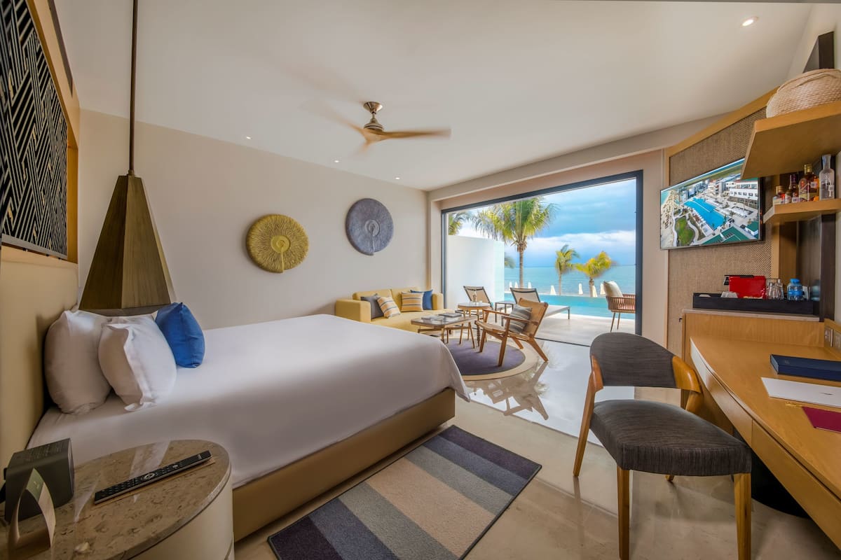 A modern hotel room with a large bed, a desk, a chair, a TV, and a balcony with a pool view.