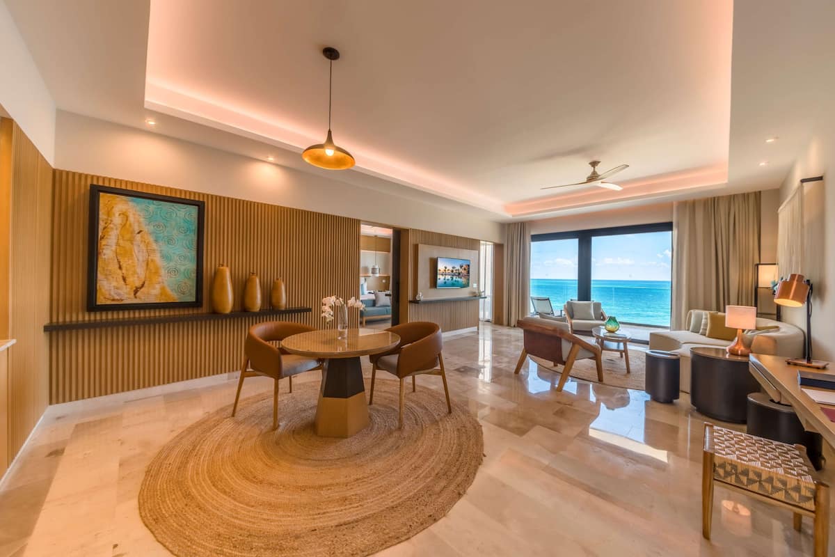 A modern hotel room with a dining area, a large window with a view of the ocean, and a ceiling fan.