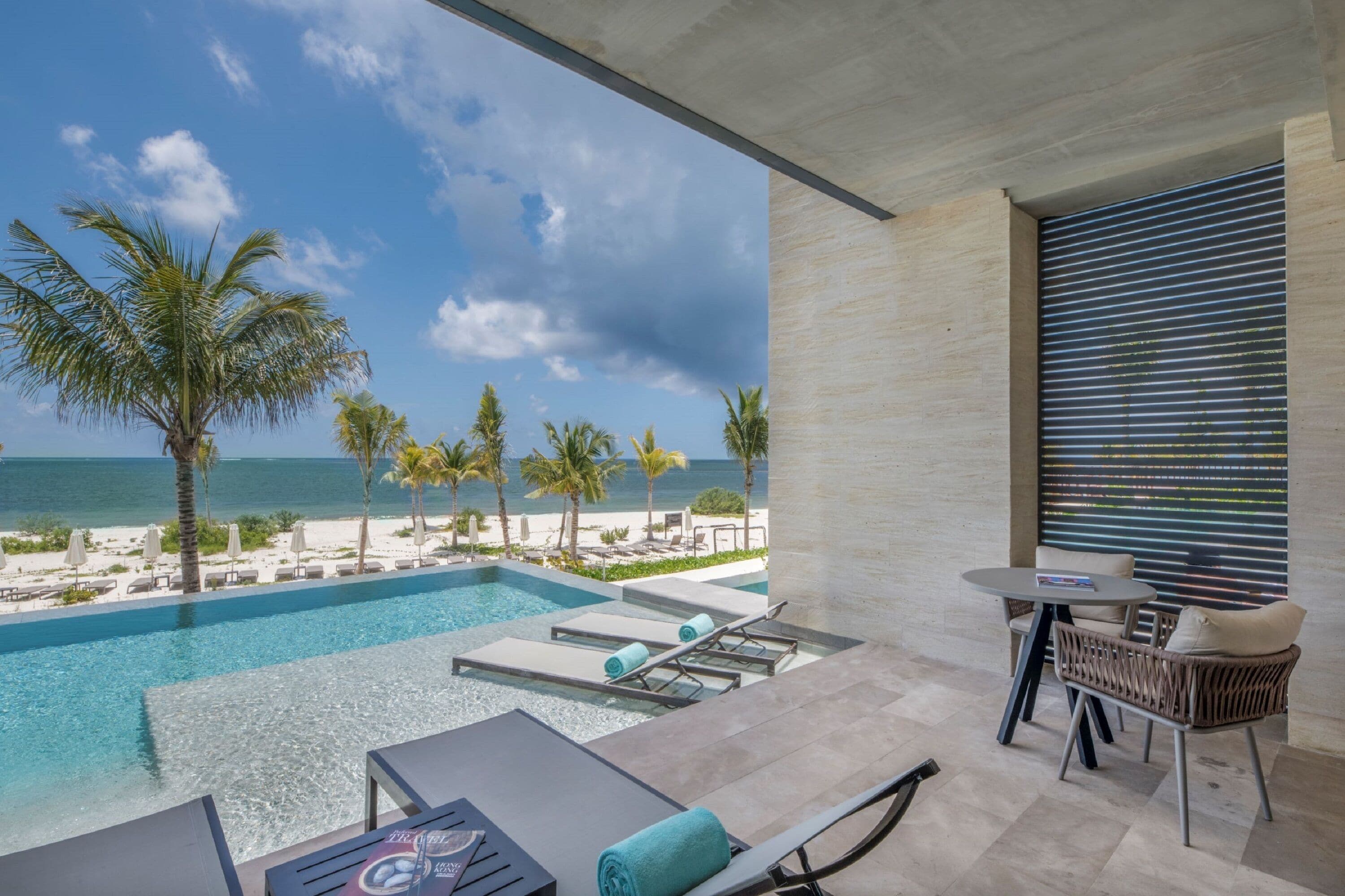 A modern pool area with a view of the beach and ocean, lounge chairs, and a table with a chair.