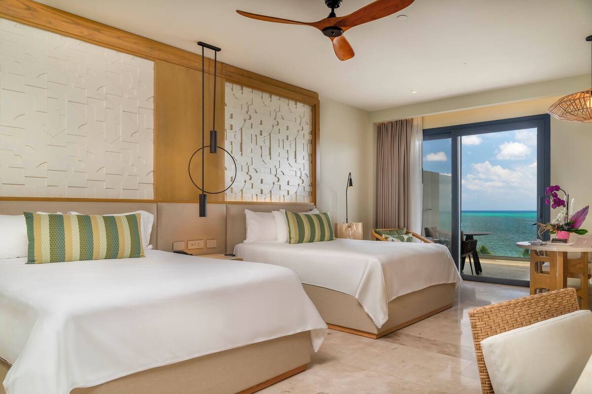 A hotel room with two beds, a ceiling fan, a view of the ocean, and a balcony with a table and chairs.