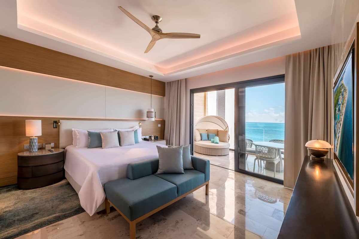 A modern hotel room with a large bed, a sofa, a ceiling fan, and a view of the ocean through the glass door.