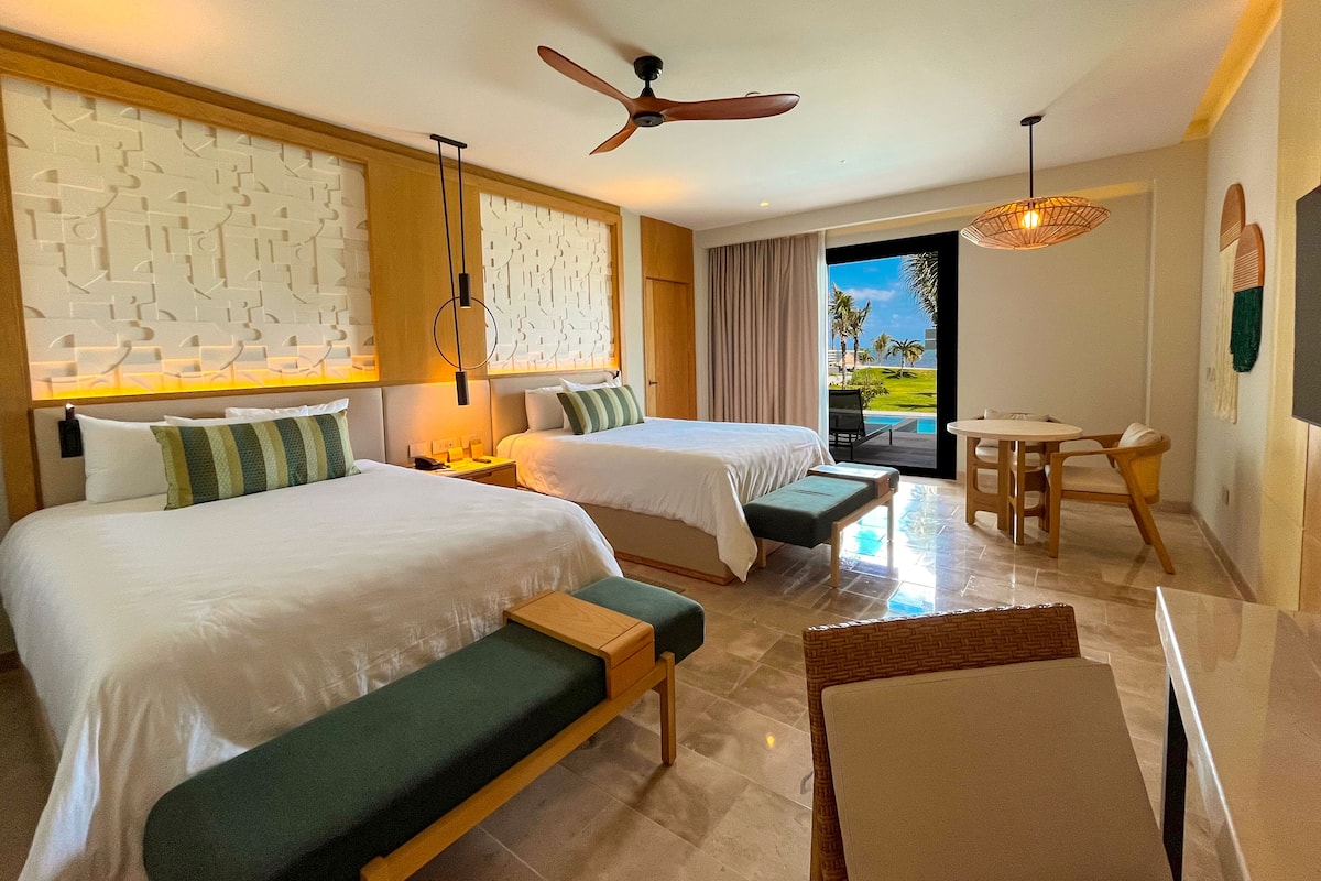 A hotel room with two beds, a desk, a chair, a ceiling fan, and a view of the outdoors.