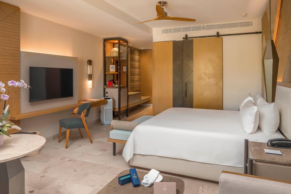 A modern hotel room with a large bed, a desk, a TV, and a seating area with a chair and a small table.