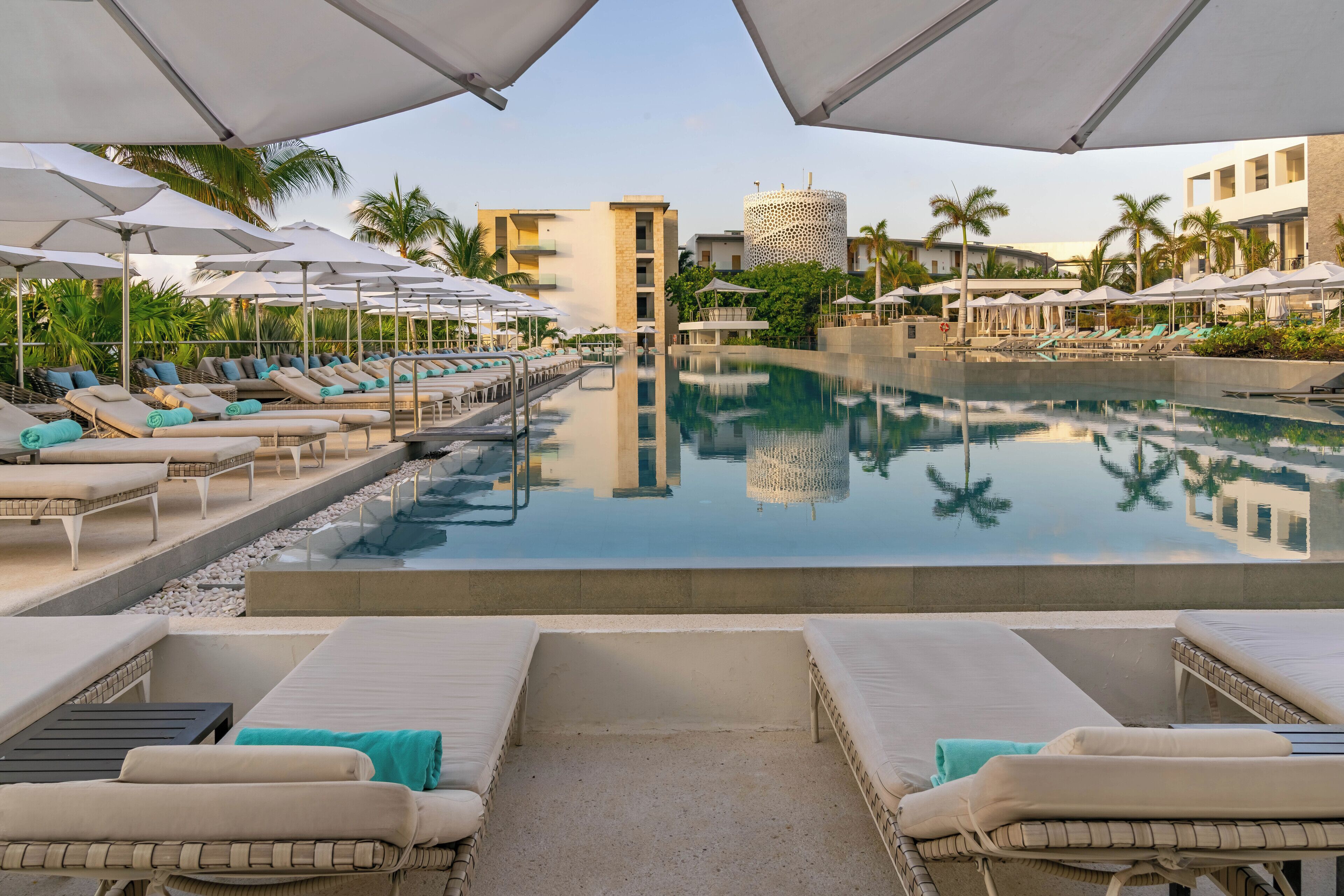 A pool area with lounge chairs and umbrellas, a clear pool, and a building with a unique facade in the background.