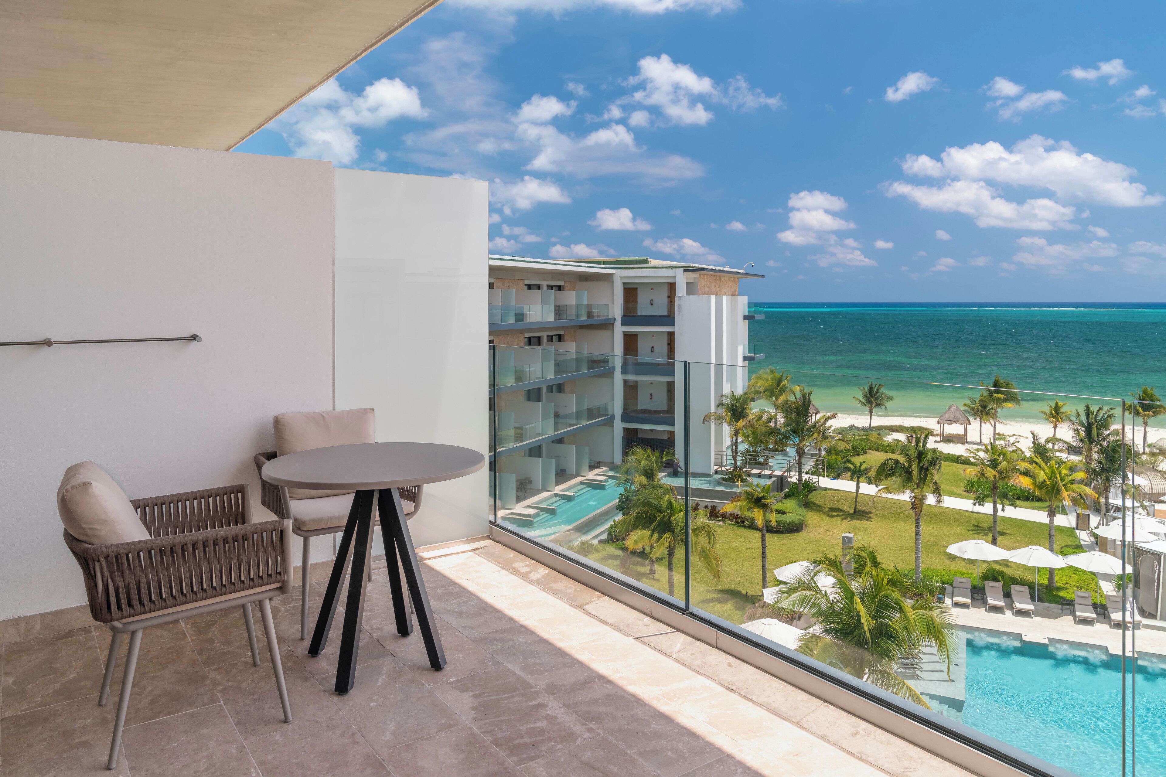 A balcony with a view of a swimming pool, lounge chairs, and a glass railing overlooking a beach and ocean.