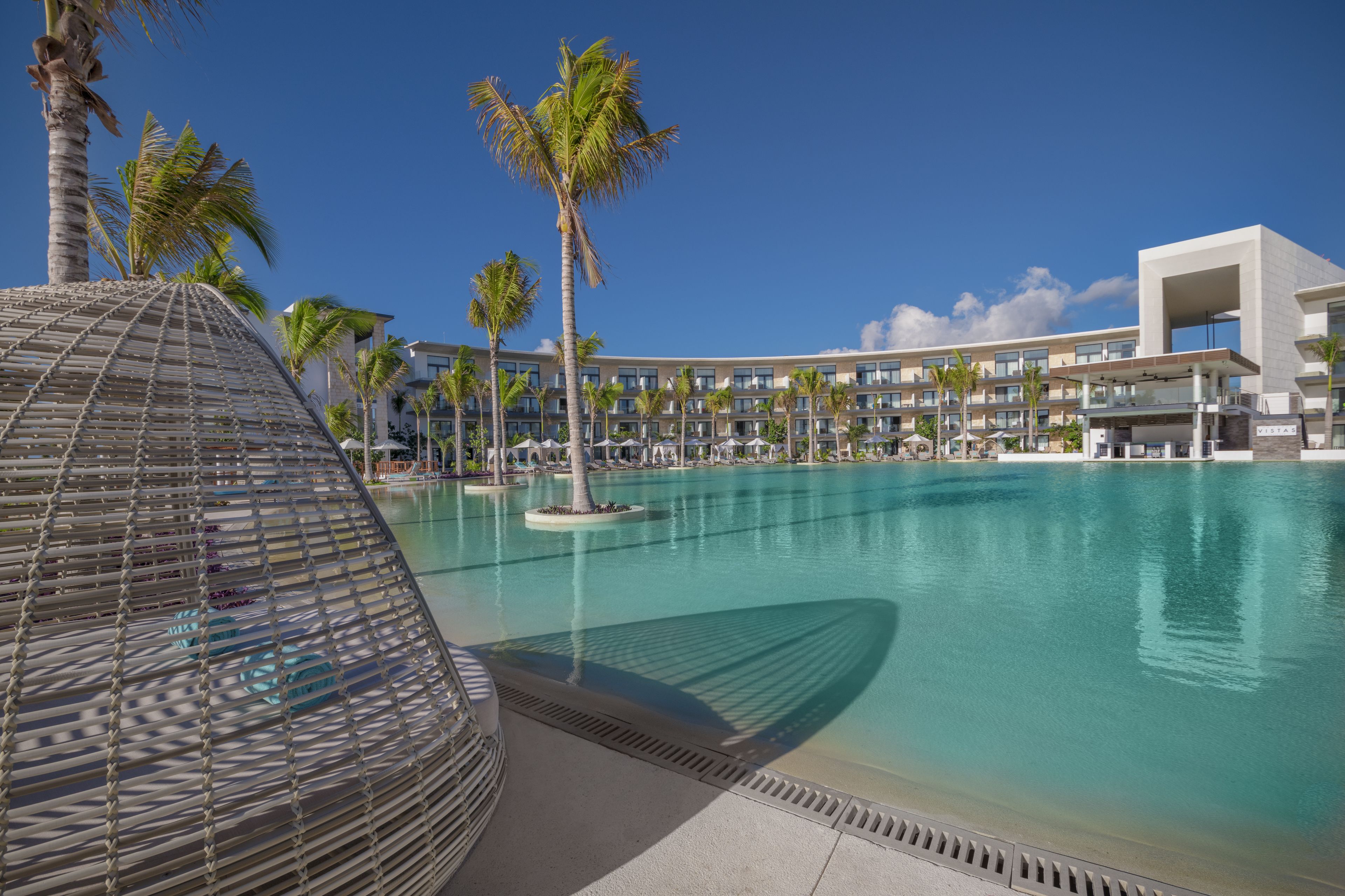 A large swimming pool with palm trees and a modern hotel building in the background.