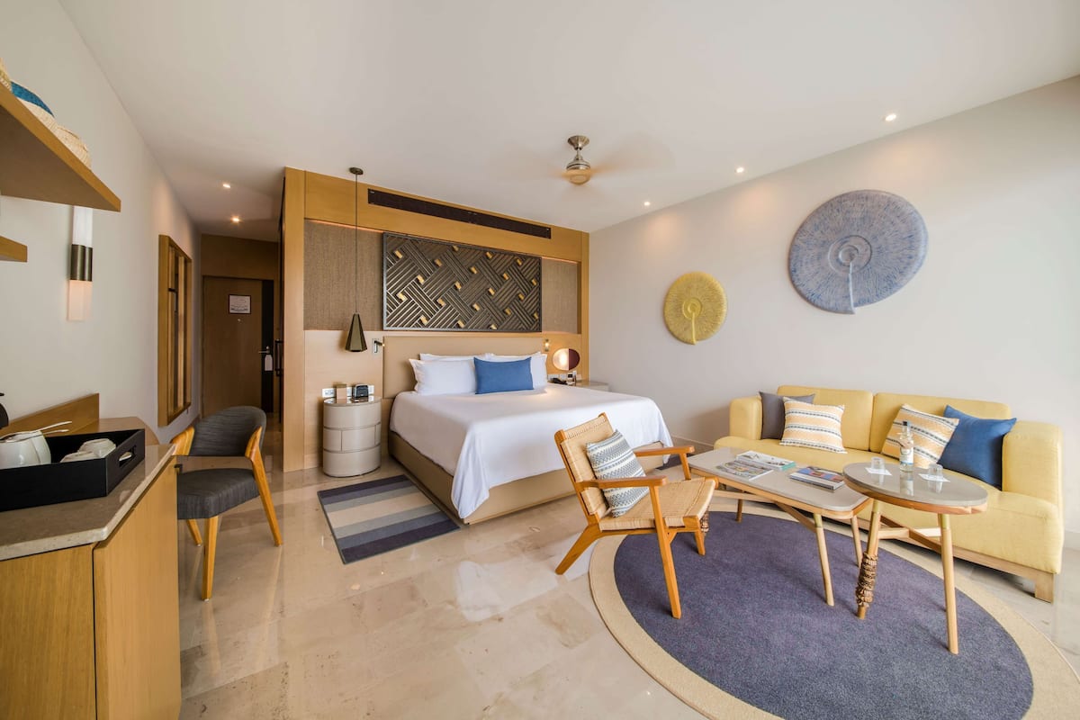 A modern hotel room with a bed, sofa, chairs, a coffee table, and a small kitchen area.