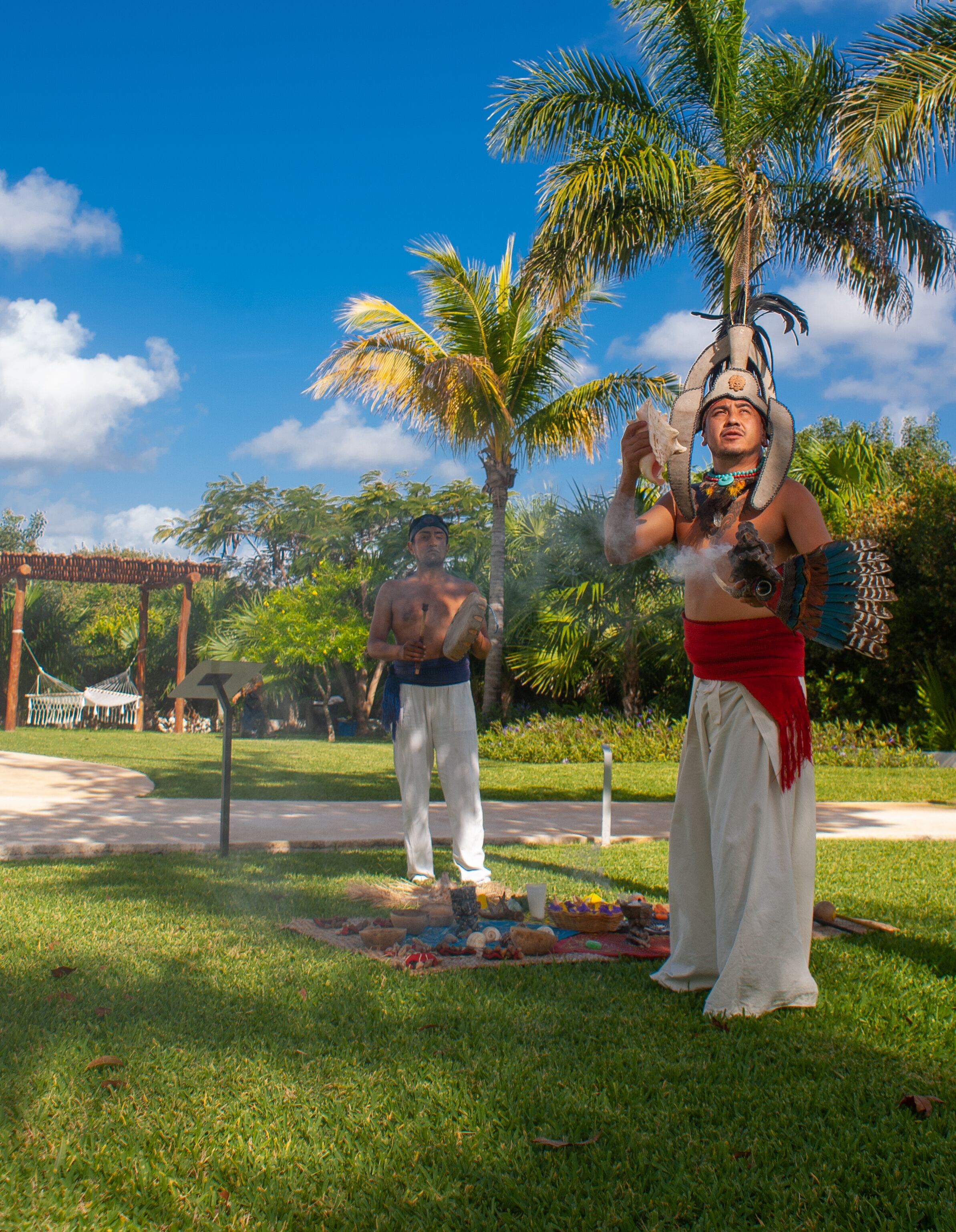 A person in traditional attire standing in a park with palm trees and a hammock in the background.
