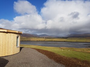 A wooden building with a gravel path leading to it, set against a backdrop of a river and distant mountains under a partly cloudy sky. - Skye Cabins (Skeabost)