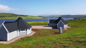 Two log cabins with black roofs, white walls, and a gravel path leading to them. They are situated on a grassy hill overlooking a lake with a wooden fence in the background. - Skye Cabins (Skeabost)