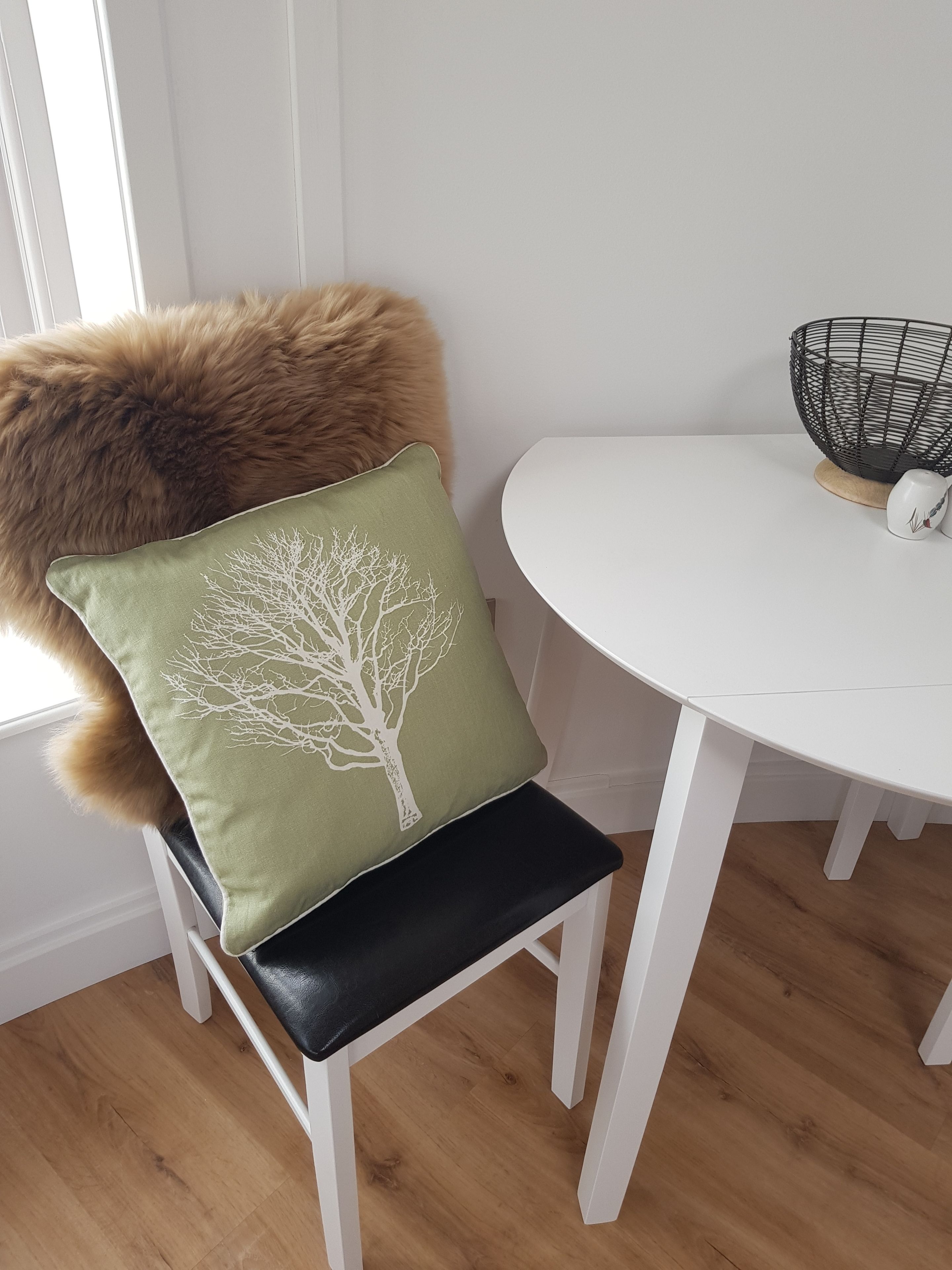A chair with a green cushion featuring a tree design, a white table with a black wire basket, and a fur throw on the chair.