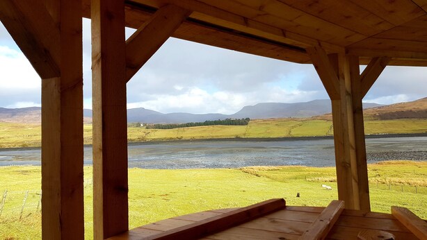 A wooden structure with a bench overlooking a river and a grassy landscape with mountains in the distance. - Skye Cabins (Skeabost)