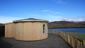 A wooden cabin with a circular shape, a door, and a window, situated by a lake with a wooden fence and hills in the background. - Skye Cabins (Skeabost)