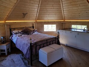 A cozy cabin interior with a wooden ceiling, a bed with a headboard, a bench, a small table, a window, and a kitchenette. - Skye Cabins (Skeabost)