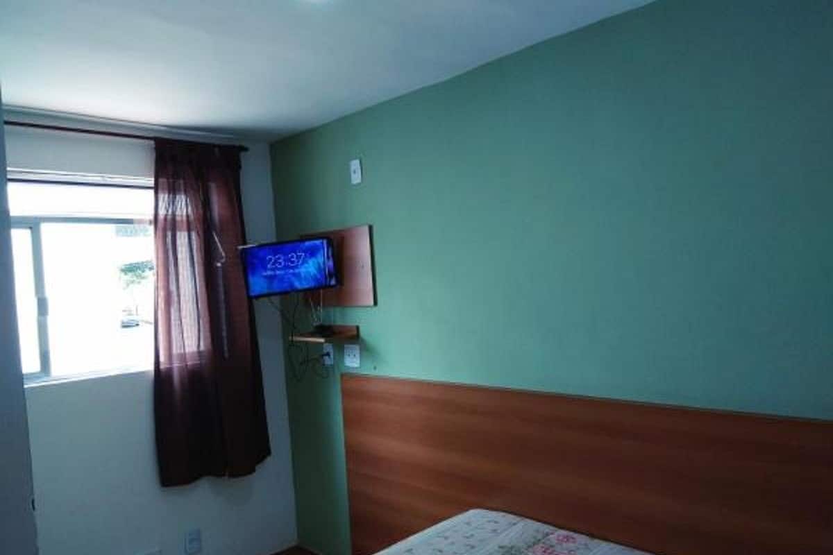 Una habitación con una cama, un televisor montado en la pared y un ventanal con cortinas.