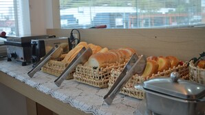 Una mesa de buffet con diversos panes y pasteles, un cortador de pan y una ventana con persianas en el fondo.