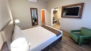 Quarto de hotel com cama, luminária de cabeceira, ar condicionado, espelho, televisão e uma cadeira verde.