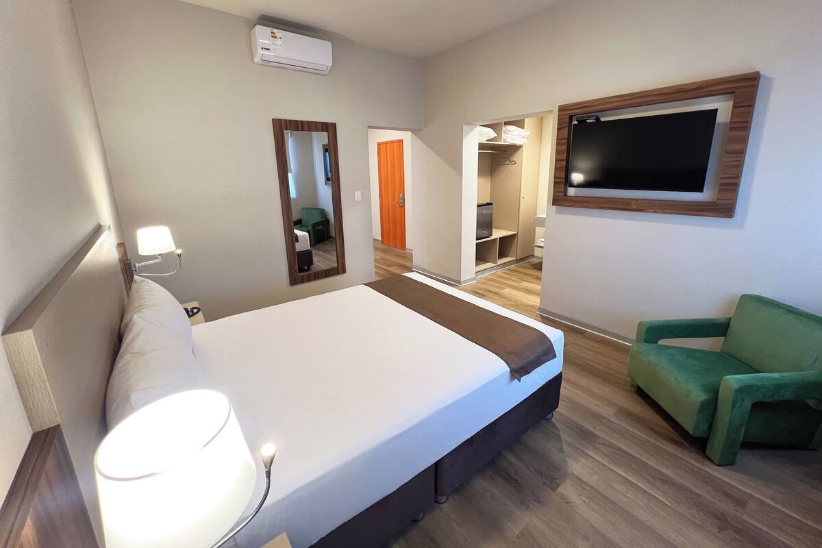 A hotel room with a bed, bedside lamp, air conditioning unit, a mirror, a television, and a green chair.