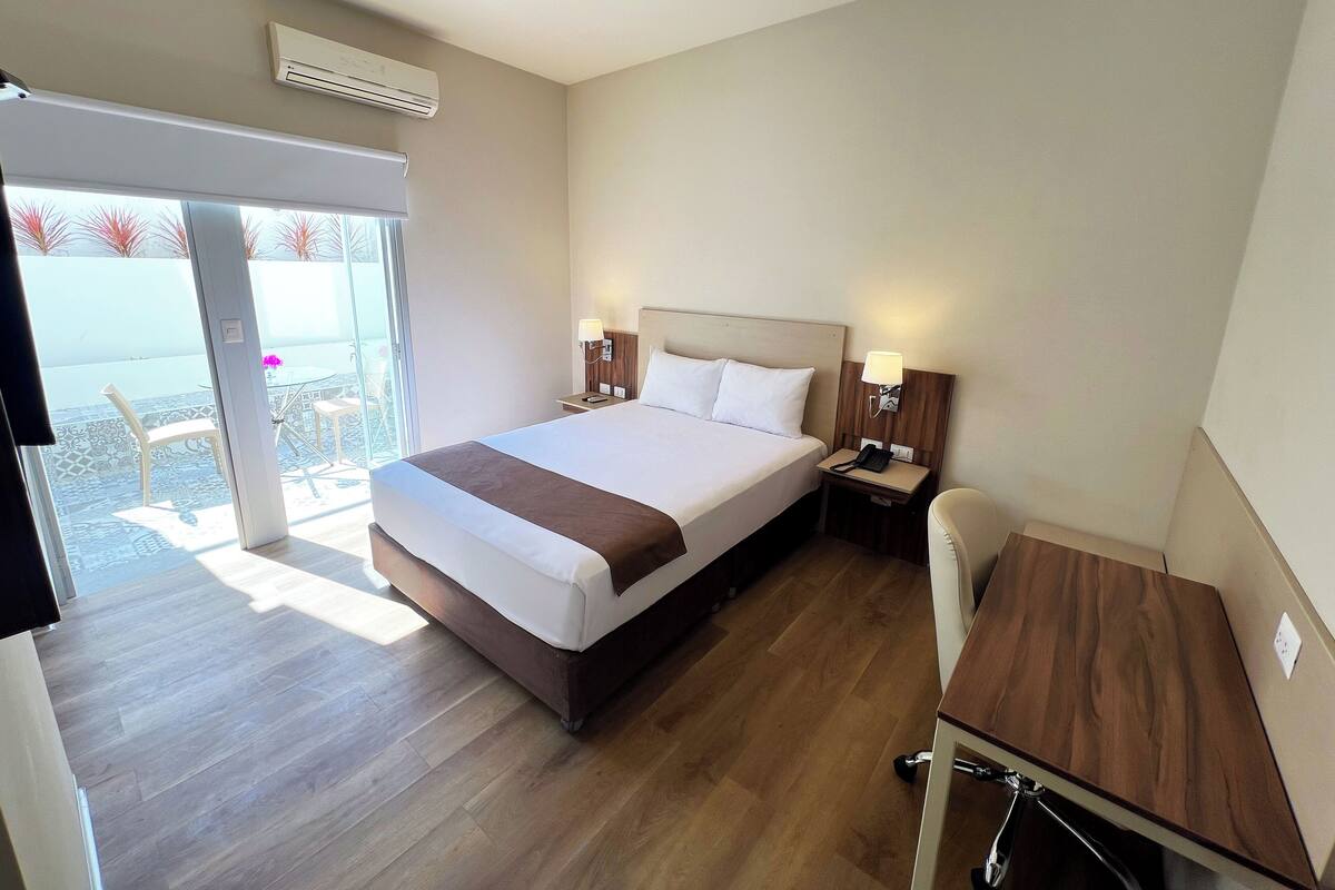 A hotel room with a bed, bedside tables, a desk, and a chair. There is a sliding glass door leading to an outdoor area with chairs and a table.