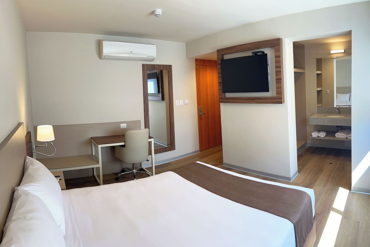 A hotel room with a bed, desk, chair, and a bathroom with a sink and mirror.