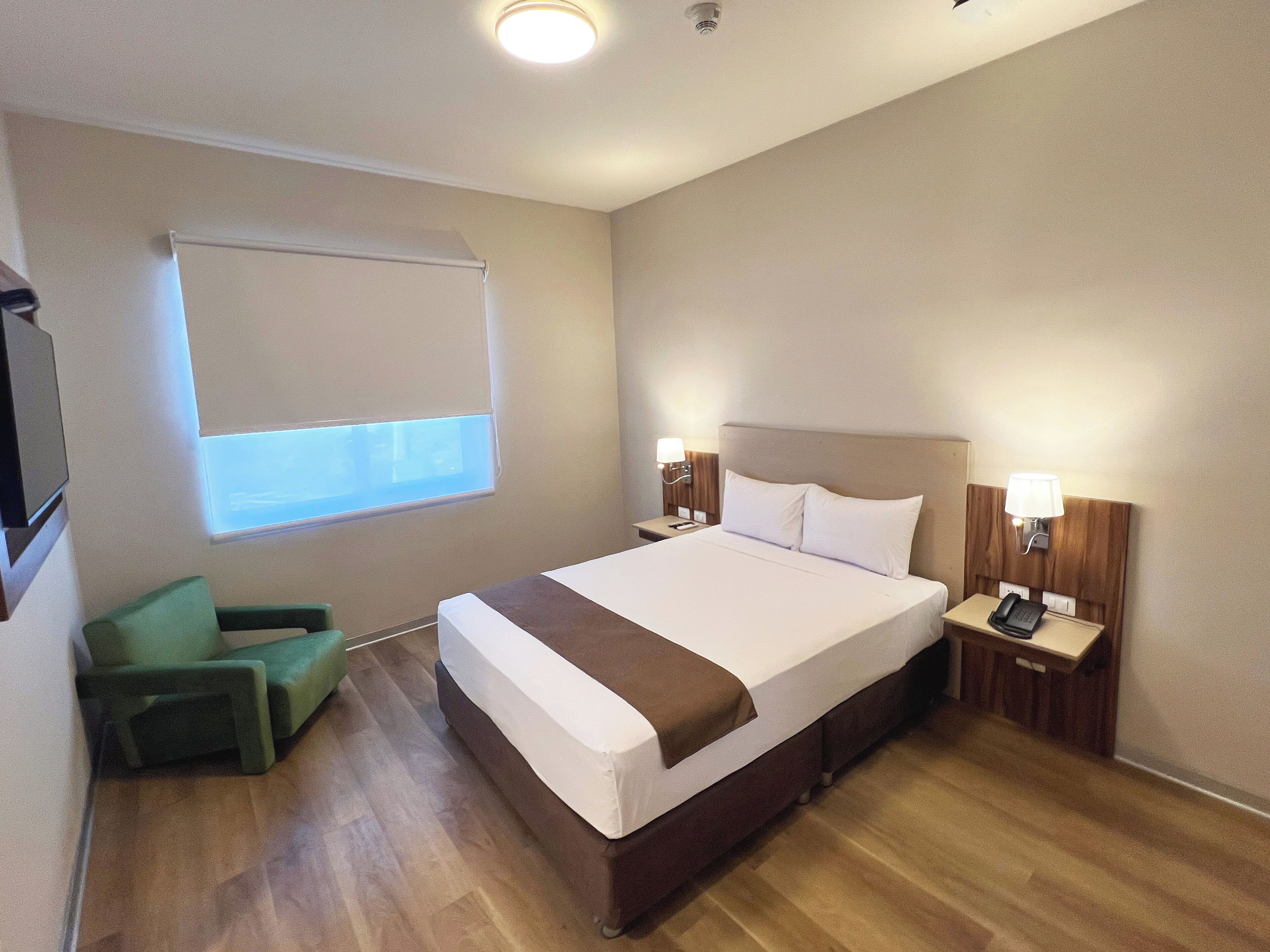 A hotel room with a large bed, bedside lamps, a desk, a chair, and a window with blinds.