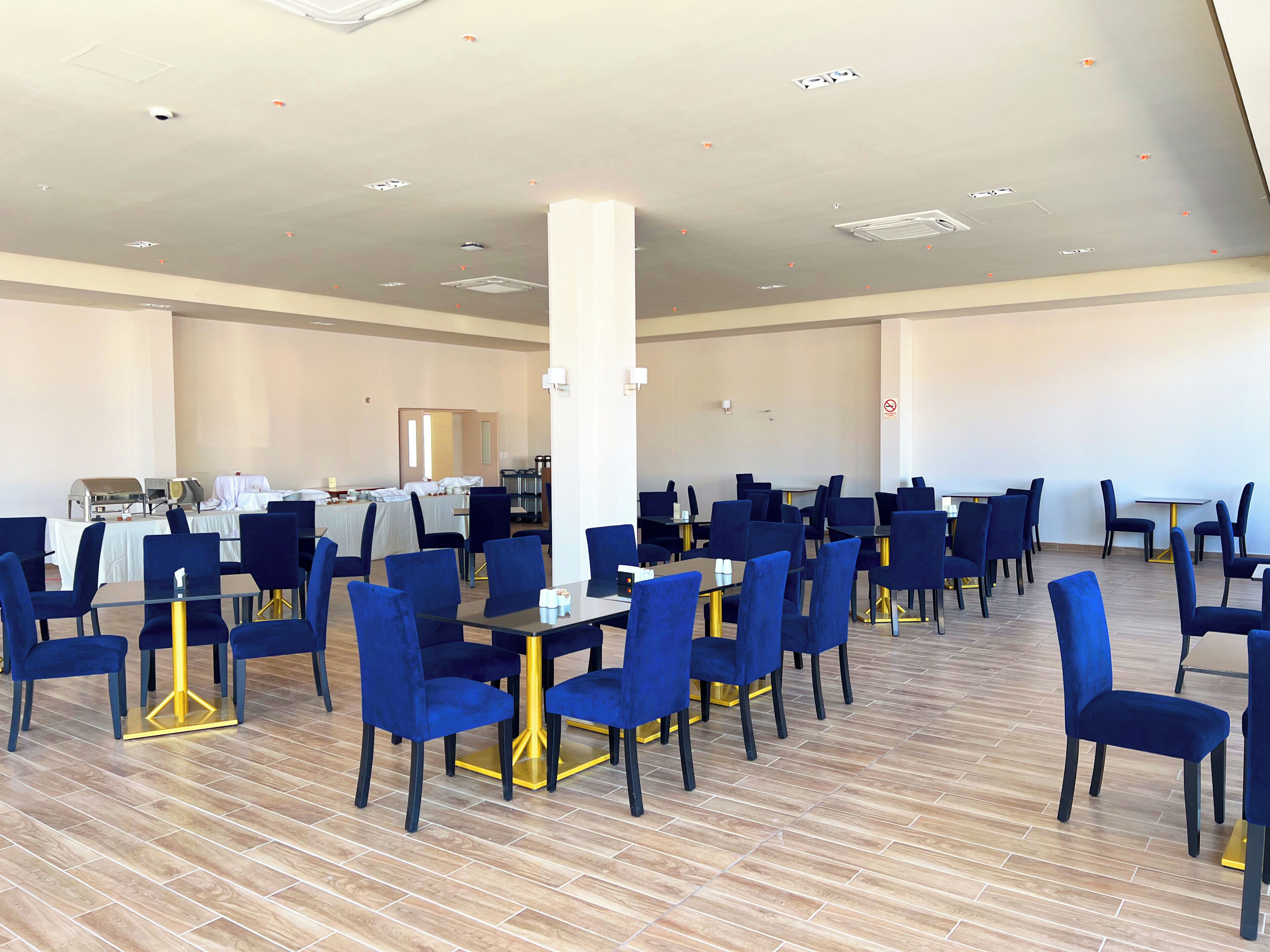 Una amplia sala de conferencias con filas de sillas y mesas azules, un techo blanco con iluminación empotrada y suelo de madera.