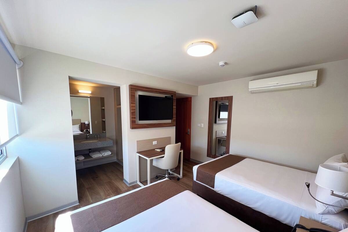A hotel room with two beds, a desk, a chair, a TV, and a bathroom visible through an open door.