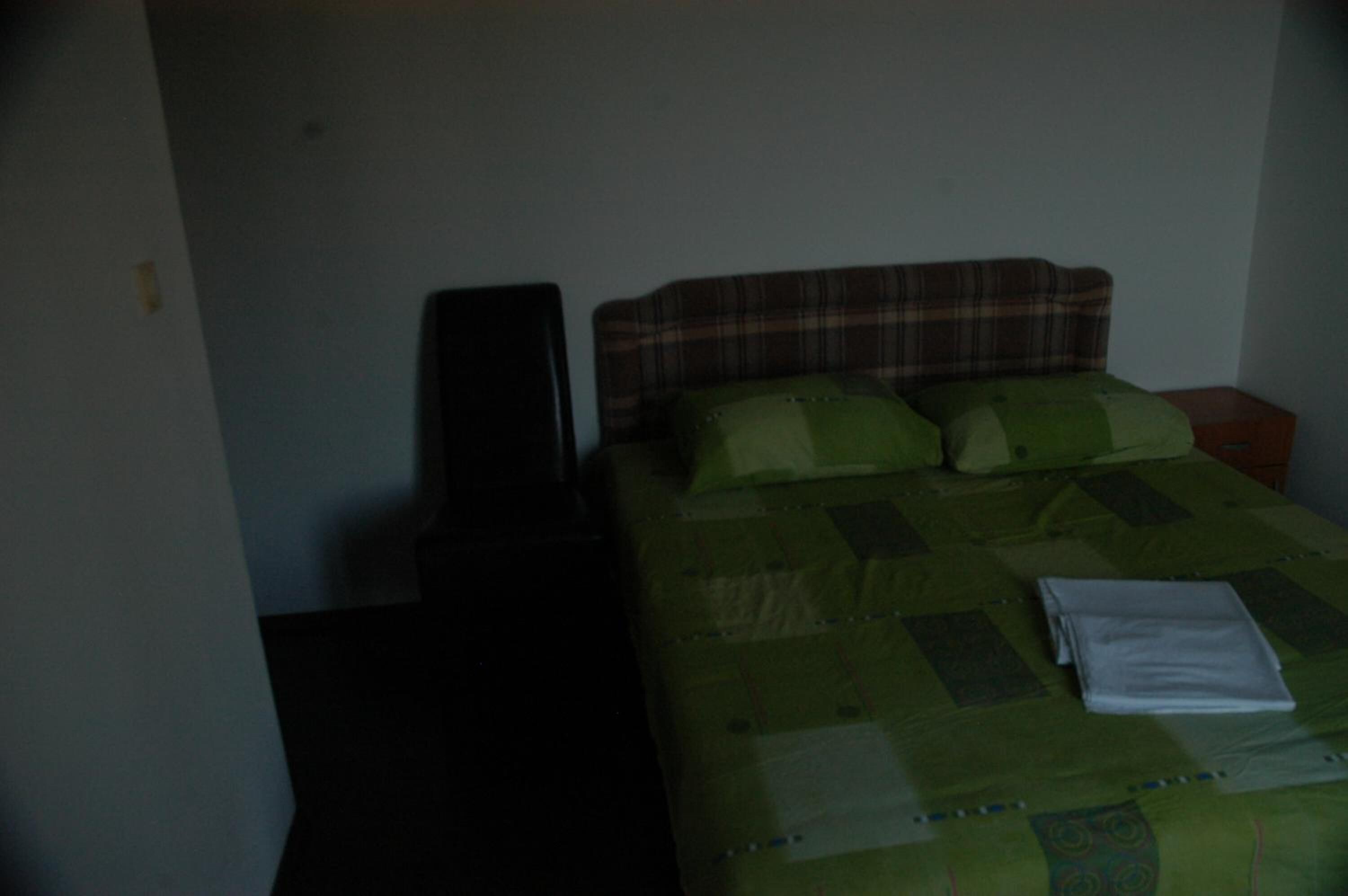 Een kleine, zwak verlichte kamer met een enkel bed, een nachtkastje en een stoel.