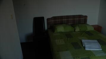 Een kleine, zwak verlichte kamer met een enkel bed, een nachtkastje en een stoel.